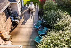 A spacious deck attached to a house, featuring a dining area and multiple chairs, surrounded by lush greenery.